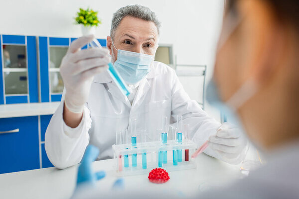 bioengineer in medical mask holding test tube near blurred colleague and coronavirus bacteria model