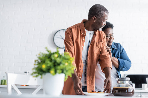 happy senior african american woman hugging husband near breakfast in kitchen 