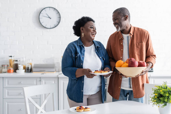 happy senior african american man holding bowl with fruits near smiling wife