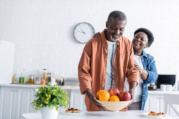 senior african american man putting bowl with fruits on breakfast table near smiling wife 