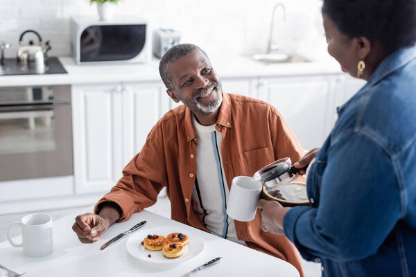 счастливый африканский американец смотрит на жену, подающую кофе во время завтрака 