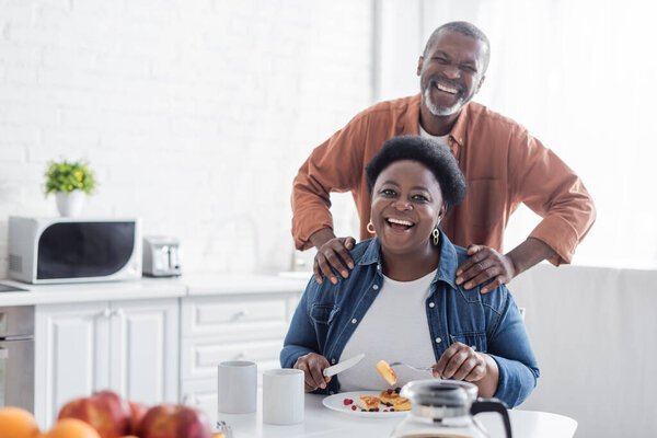 happy and senior african american man standing behind wife during breakfast 