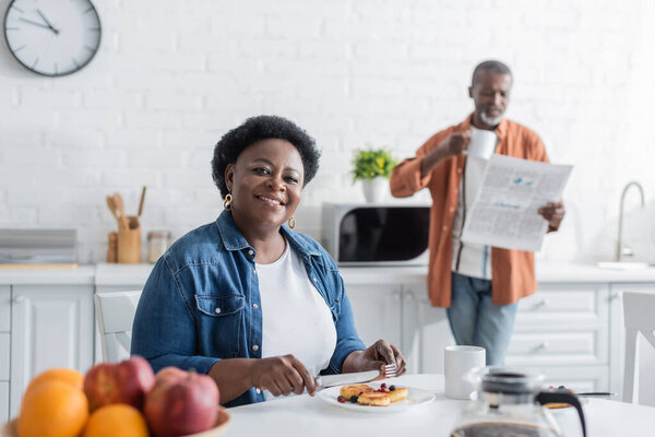 happy and senior african american woman having breakfast near blurred husband with newspaper
