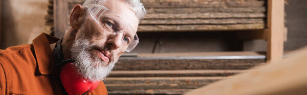 bearded furniture designer in goggles working in woodwork studio, banner