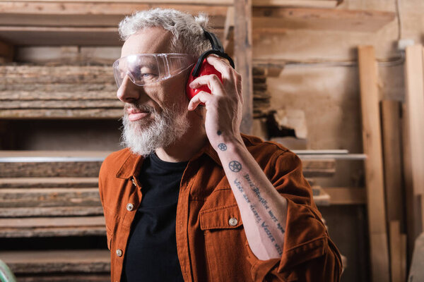tattooed carpenter in goggles adjusting protective earmuffs in workshop