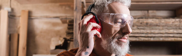 bearded carpenter in goggles adjusting protective earmuffs, banner