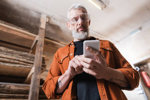 low angle view of bearded furniture designer using smartphone in woodwork studio
