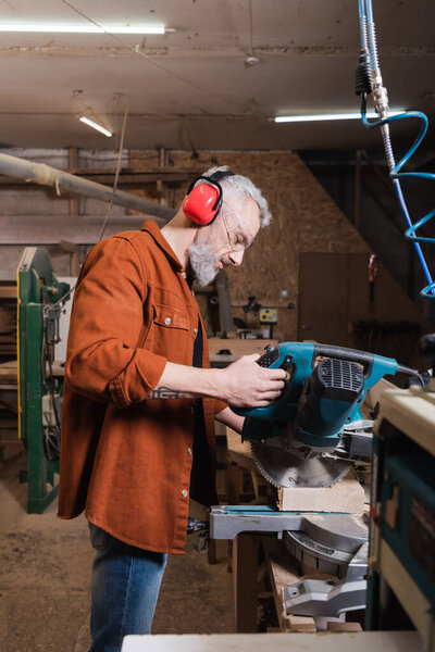 side view of carpenter in protective earmuffs cutting timber on miter saw