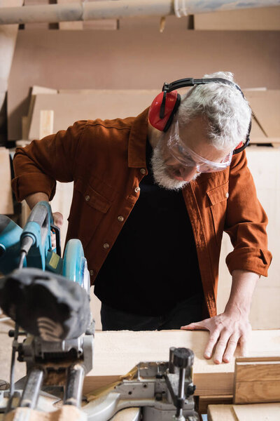 grey haired woodworker cutting timber on miter saw in carpentry studio