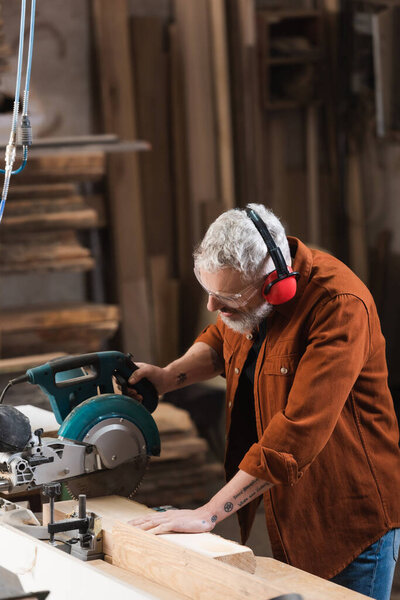 tattooed carpenter cutting wood with miter saw in woodwork studio