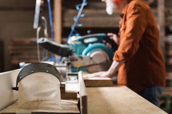 selective focus of face shield near cropped carpenter working on blurred miter saw