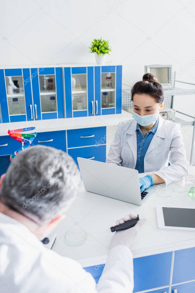 scientist in medical mask working on laptop near digital tablet with blank screen and blurred colleague