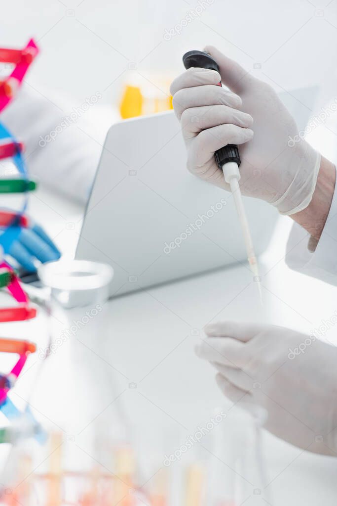 cropped view of geneticist in latex gloves holding micropipette near blurred laptop