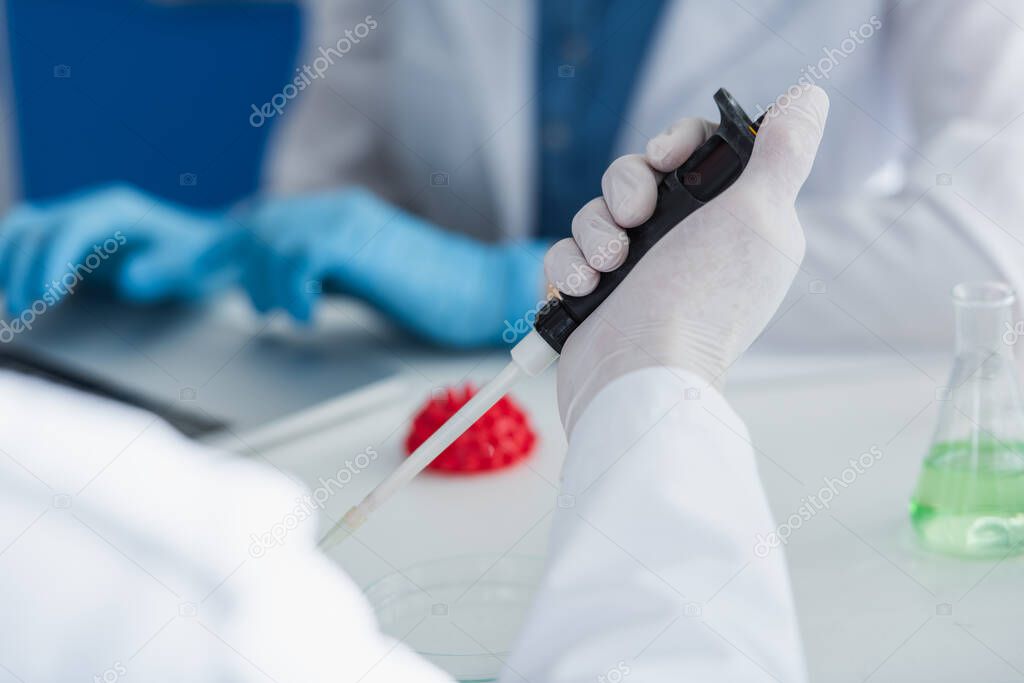 cropped view of scientist in latex glove holding micropipette near blurred model of coronavirus bacteria