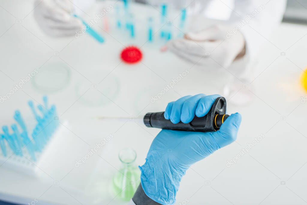 cropped view of scientist in latex glove holding micropipette near blurred colleague