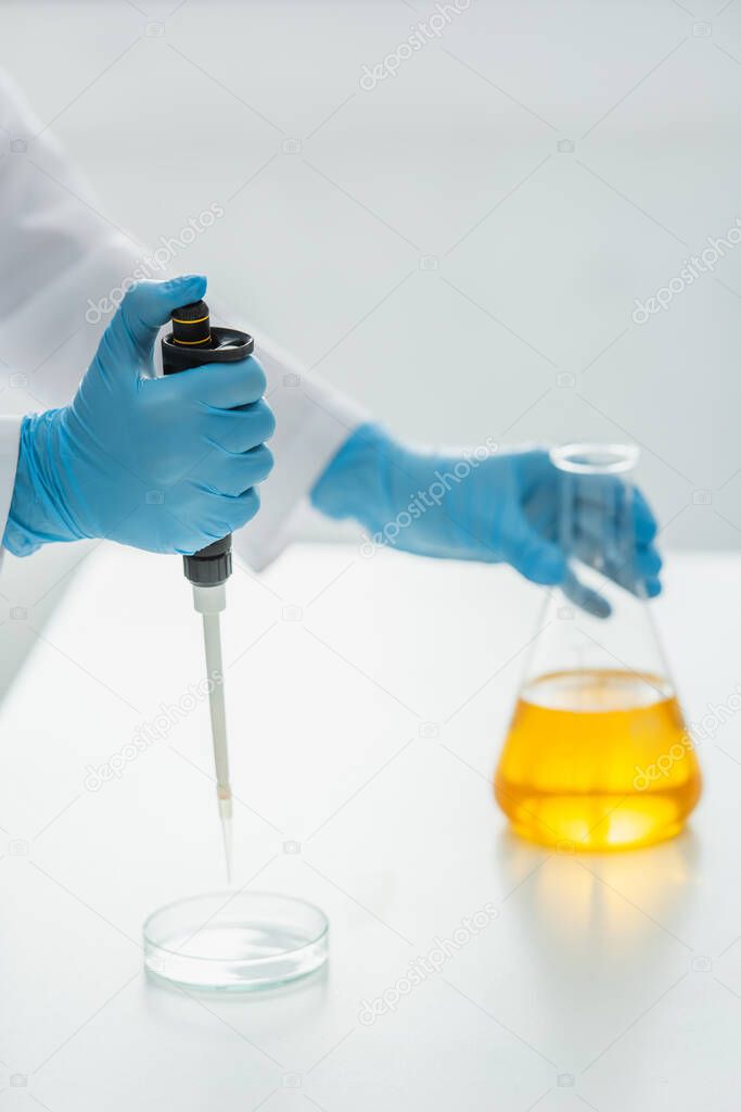 cropped view of scientist with micropipette near petri dish and flask with yellow liquid