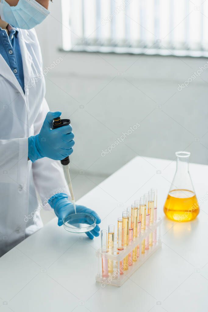 partial view of woman in medical mask and white coat working with micropipette and petri dish