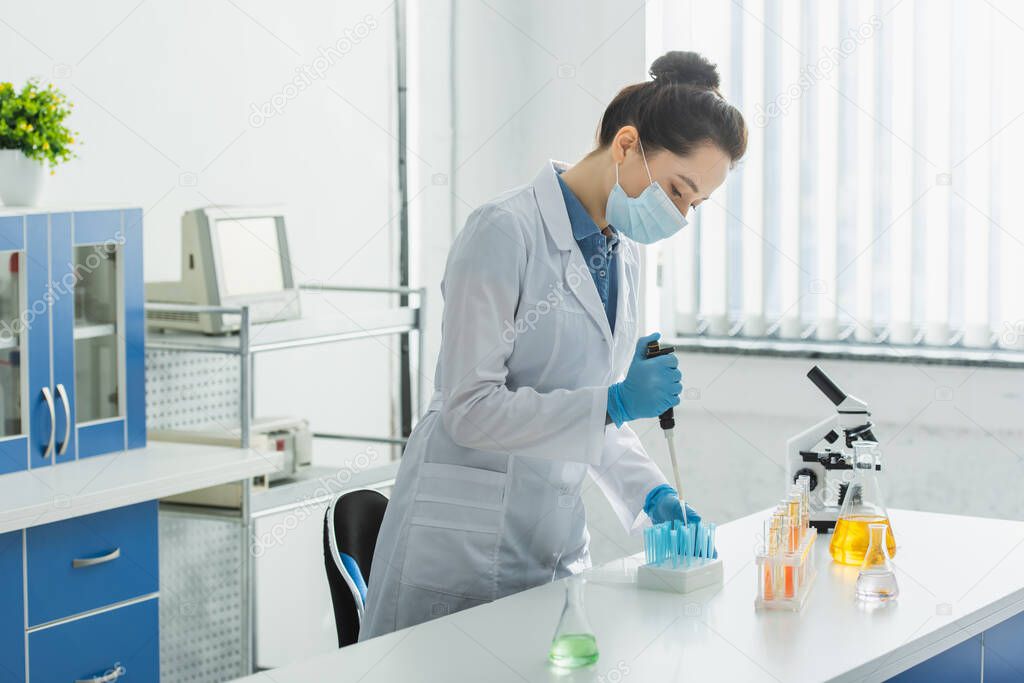 bioengineer in medical mask and white coat working with micropipette and test tubes near microscope