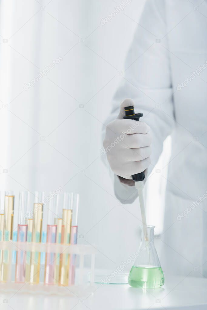 partial view of geneticist in latex glove holding micropipette near flask and test tubes