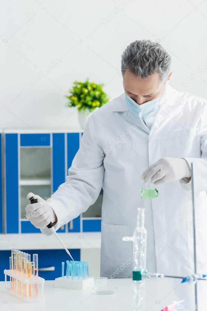 bioengineer in medical mask holding micropipette and flask while working with test tubes in lab