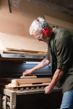 side view of grey haired carpenter polishing board in sander machine clipart