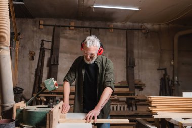 Mutlu orta yaşlı marangoz atölyede ortak makine üzerinde çalışıyor.