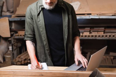 cropped view of furniture designer writing in notebook near laptop on workbench clipart