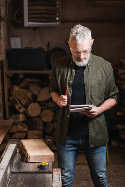 bearded furniture designer in goggles standing with notebook and pencil in workshop
