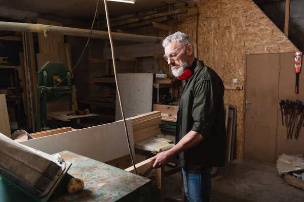 bearded carpenter holding wooden plank near thickness planer in workshop