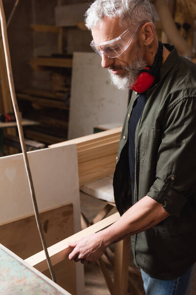 mature carpenter in goggles holding wooden plank in woodwork studio