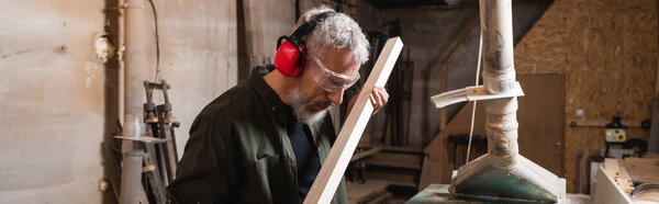 carpenter in earmuffs and goggles checking wooden plank in workshop, banner