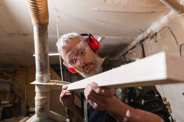 woodworker in googles and protective earmuffs checking plank in workshop