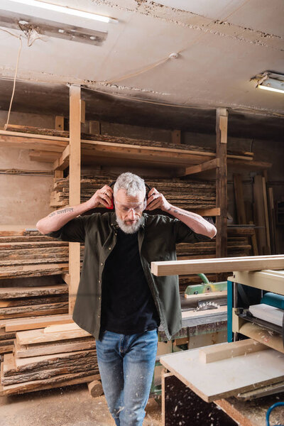 bearded carpenter adjusting protective earmuffs near thickness planer