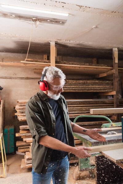 carpenter in protective earmuffs pointing at plank near sawdust 