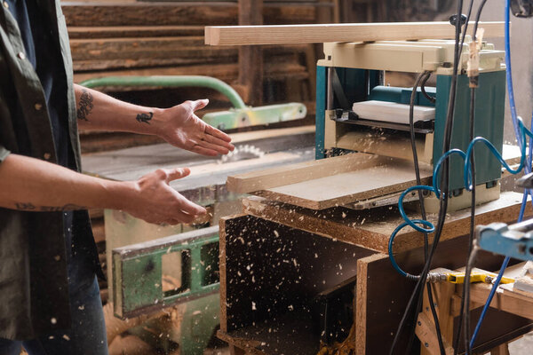 partial view of tattooed furniture designer pointing at boards in thickness planer 