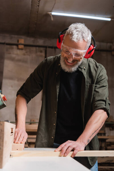 smiling carpenter in goggles working on jointer machine in workshop