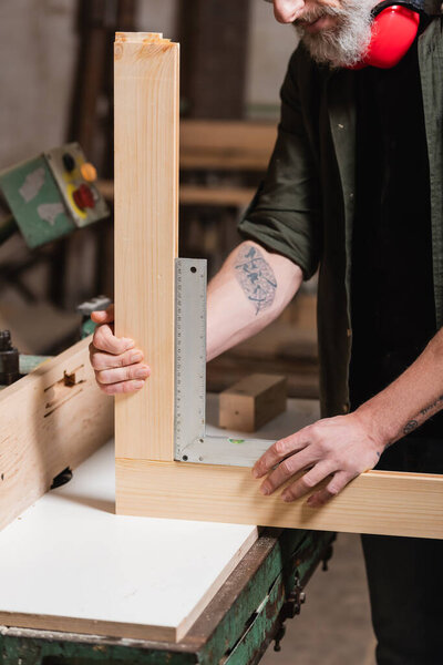 cropped view of tattooed joiner measuring wooden details with corner ruler
