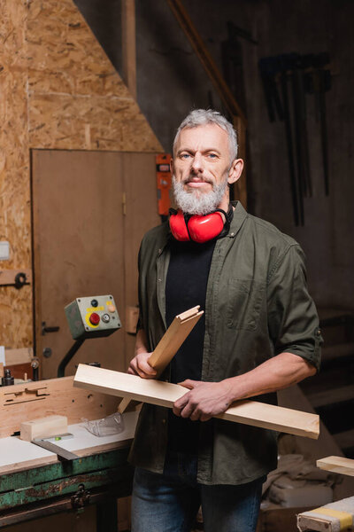 bearded carpenter holding wooden details and looking at camera in workshop