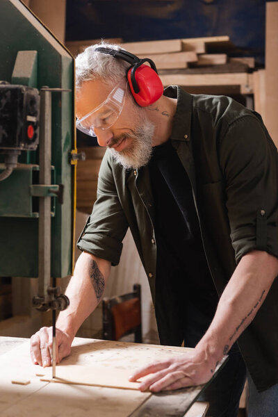 bearded carpenter in goggles cutting plywood with band saw