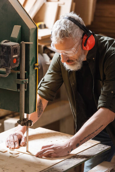 bearded woodworker in goggles cutting plank on band saw