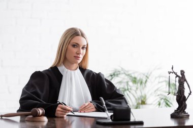 blonde judge looking at camera while sitting with pen near gavel and blurred themis statue clipart