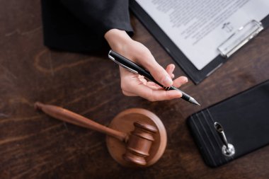 top view of cropped prosecutor holding pen near gavel and clipboard on wooden desk clipart
