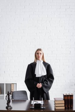 prosecutor in black mantle looking at camera near desk and white wall in office clipart