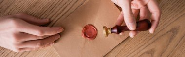 partial view of notary holding stamper near envelope with wax seal on wooden desk, banner clipart