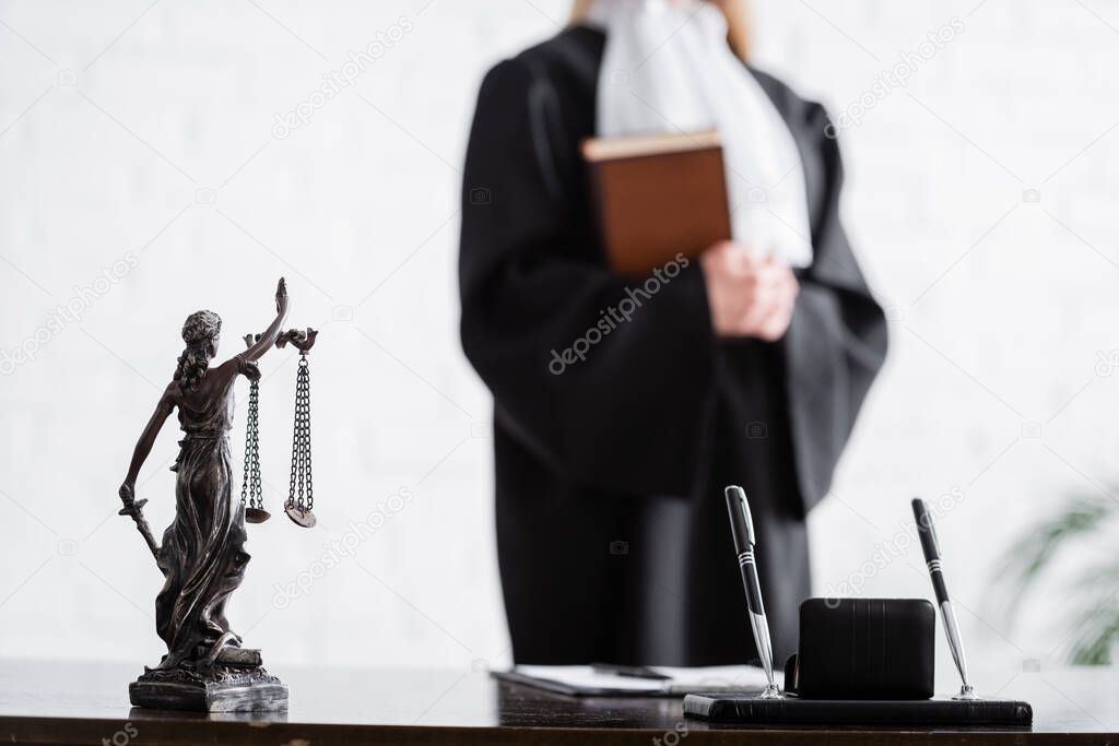selective focus of themis statue and pens near cropped prosecutor with book on blurred background