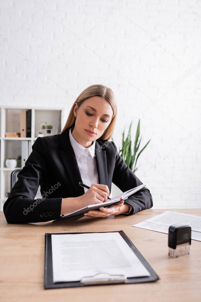blonde lawyer writing in notebook near documents and stamper on desk