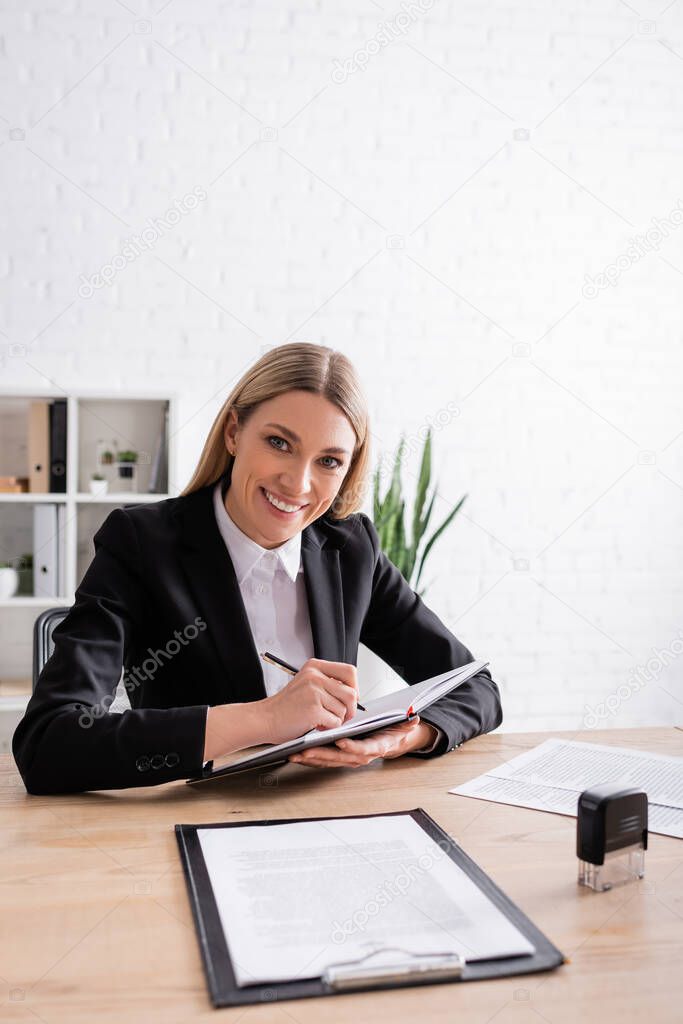 cheerful notary looking at camera while writing in notebook at workplace