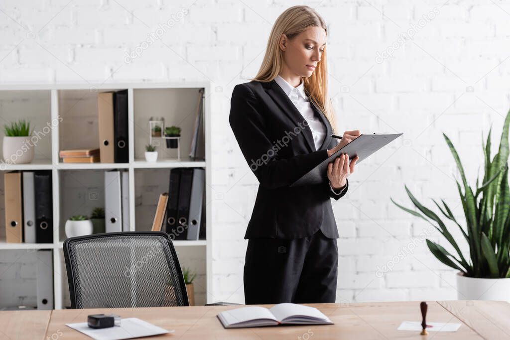 blonde lawyer writing on clipboard while standing in office