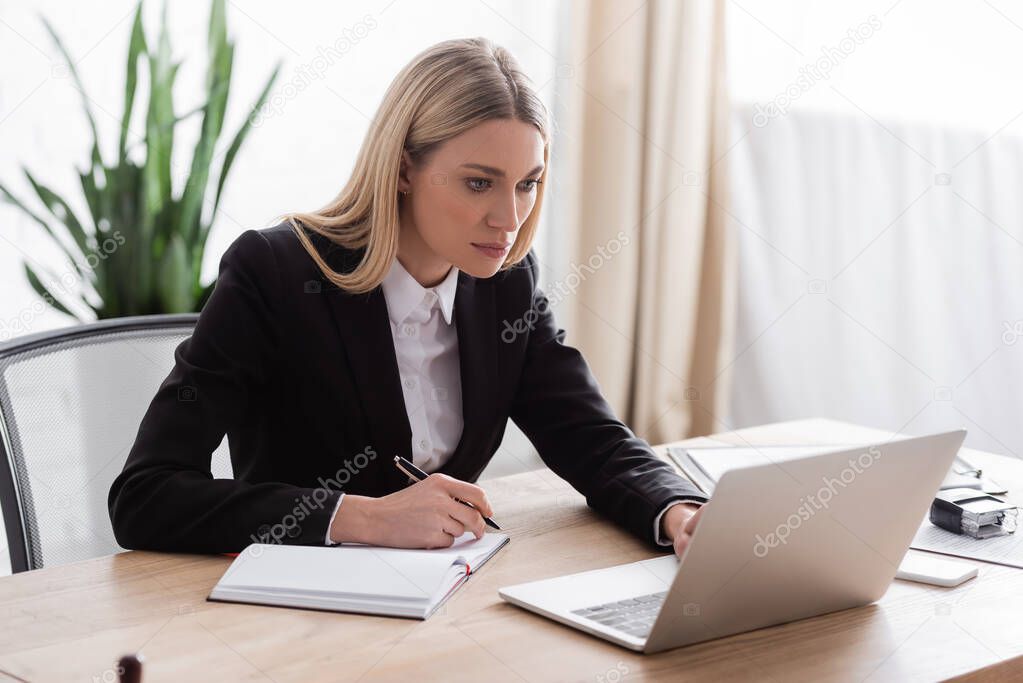 lawyer working with laptop while holding pen near blank notebook