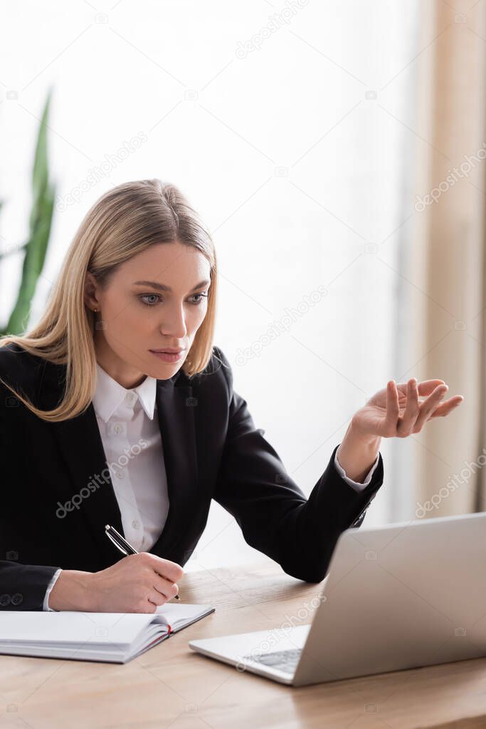 blonde notary gesturing near laptop while holding pen near notebook